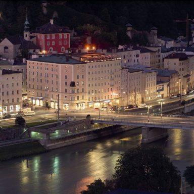 Nachtansicht einer Stadt mit beleuchteten Gebäuden und einem Fluss im Vordergrund.