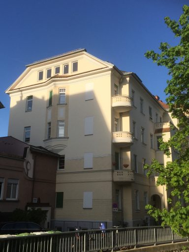 Helles mehrstöckiges Gebäude mit Balkonen, umgeben von Bäumen und klarem Himmel.